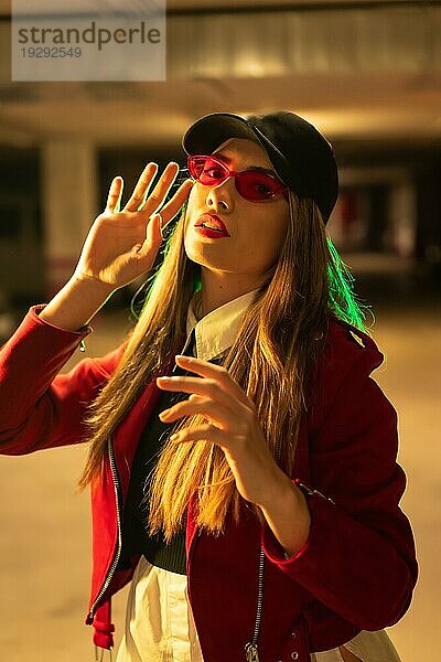 Fotografie mit roten und grünen Neons auf einem Parkplatz. Junge hübsche blonde kaukasische Frau in einem roten Anzug  Sonnenbrille und einer schwarzen Kappe