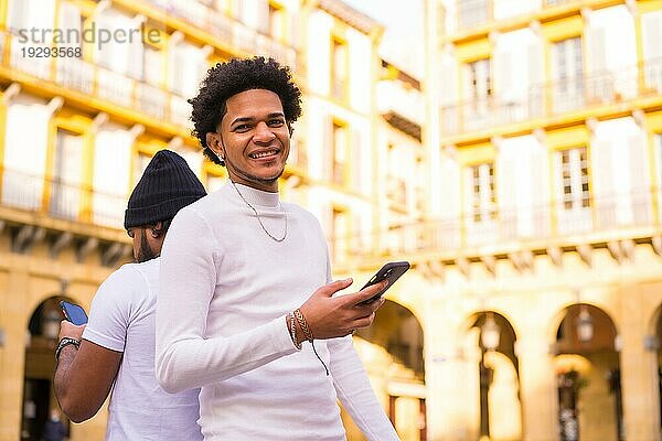Lifestyle  zwei schwarze Latino Freunde  die Nachrichten versenden oder soziale Netzwerke auf ihrem Handy beobachten. Junge mit Afrohaar und Junge mit Hut und Kopfhörern