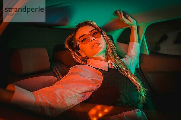 Stadtfotografie mit roten und grünen Neons auf dem Rücksitz eines Autos einer jungen blonden kaukasischen Frau mit Herzbrille