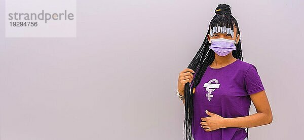 Internationaler Frauentag im Jahr der Coronaviruspandemie Covid 19. Dominikanische Frau mit feministischem lila Hemd und Gesichtsmaske  grauer einfarbiger Hintergrund mit Kopier und Einfügefläche
