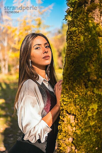 Ein junges  kaukasisches  brünettes Collegemädchen in einem Park neben einem Baum an einem Herbstnachmittag in einer modischen Pose