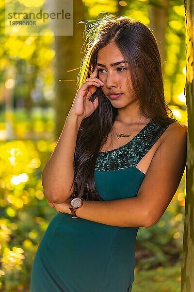 Eine junge brünette Latina mit langen glatten Haaren lehnt in einem grünen Kleid an einem Baum. Porträt mit einem süßen und sinnlichen Blick  vertikales Foto