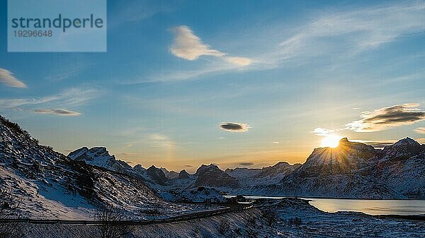 Blick auf die untergehende Sonne hinter einem schneebedeckten Berg mit Straße und See auf den Lofoten in Norwegen