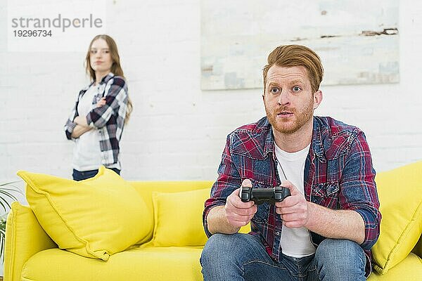 Ernster junger Mann  der ein Spiel mit einem Videoregler spielt  während seine Freundin im Hintergrund steht