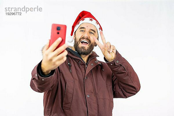 Sehr glücklicher junger kaukasischer Mann mit roter Weihnachtsmütze  der ein Selfie auf einem weißen Hintergrund macht