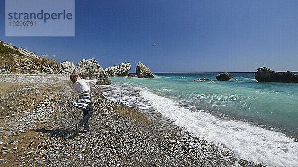 Junge schleudert einen Stein in die Luft Richtung Meer  Strand  Südküste  grünes Meer  blauer wolkenloser Himmel  Brandung  Felsen im Meer  Nähe Mirtos  Provinz Lassithi  Kreta  Griechenland  Europa