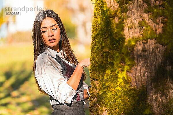 Eine junge kaukasische  brünette Studentin  die an einem Herbstnachmittag in einem Park neben einem Baum sitzt