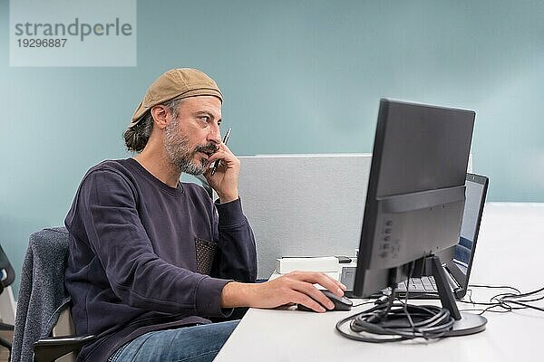 Moderner und stilvoller reifer Mann  der mit dem Telefon spricht  während er einen Computer in einer Arbeitsgruppe benutzt