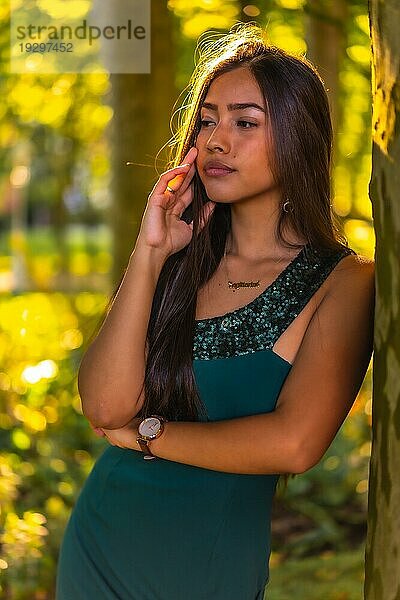 Eine junge brünette Latina mit langen glatten Haaren lehnt in einem grünen Kleid an einem Baum. Porträt mit einem süßen und sinnlichen Blick  vertikales Foto