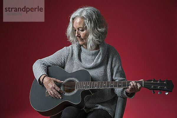 Porträt ältere Frau sitzend Stuhl spielen Gitarre gegen roten Hintergrund