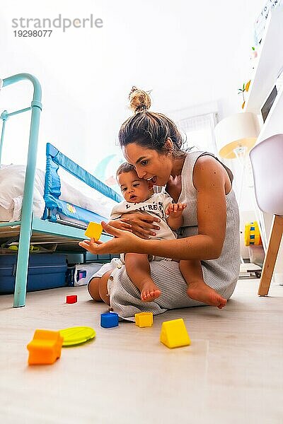 Junge kaukasische Mutter spielt mit ihr im Zimmer mit Spielzeug. Baby weniger als ein Jahr lernen die ersten Lektionen von ihrer Mutter. Mutter  die mit ihrem Sohn spielt und ihn liebevoll umarmt