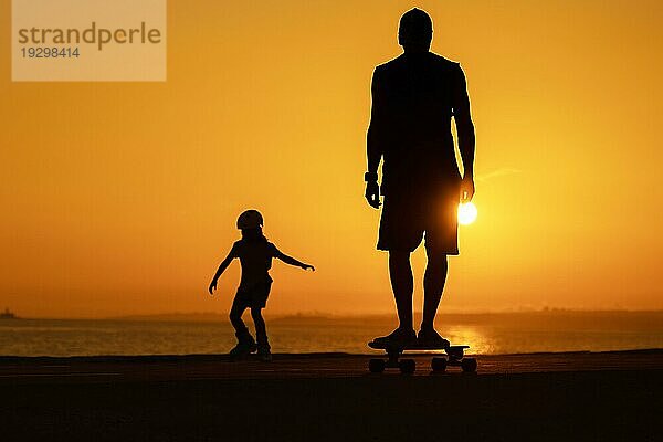 Silhouette eines Mannes und eines Kindes  die bei strahlendem Sonnenuntergang auf Skateboards und Rollschuhen skaten. Mittlere Aufnahme