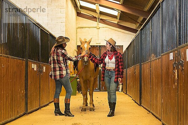 Zwei Cowgirl Frauen haben Spaß und lachen mit einem Pferd in einem Stall  amerikanische Cowboyhüte  karierte Hemden und Jeans