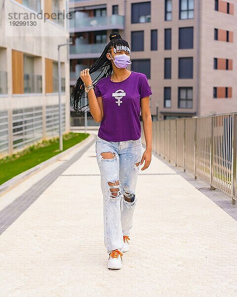 Internationaler Frauentag im Jahr der Coronaviruspandemie Covid 19. Dominikanische Frau mit feministischem lila Hemd und Gesichtsmaske beim Spaziergang durch die Stadt