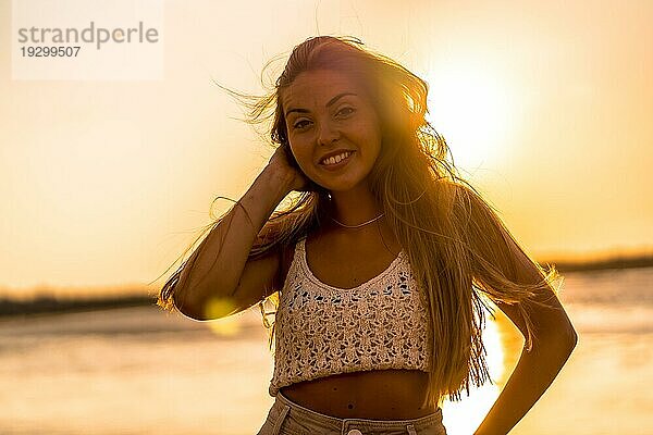 Sommerlicher Lebensstil. Eine junge blonde kaukasische Frau in einem weißen kurzen Wollpullover am Strand bei Sonnenuntergang. Lächelnd und Blick in die Kamera