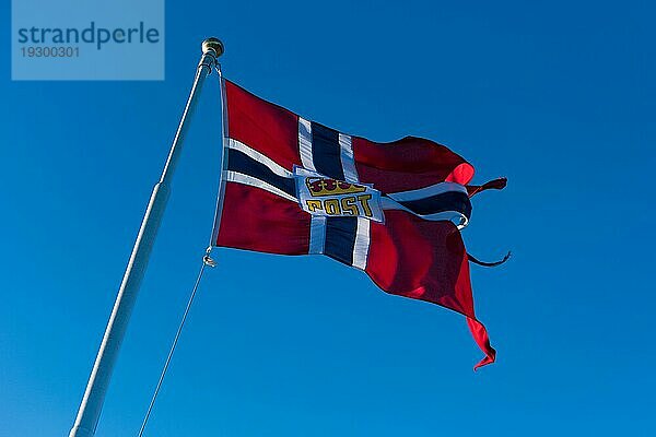 Norwegische Postflagge  gehisst am Mast eines Postbootes  das im Wind weht  mit blauem Himmel im Hintergrund