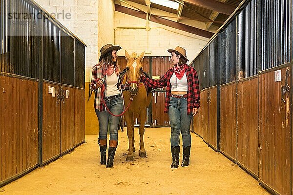 Zwei Cowgirl Frauen haben Spaß und lachen mit einem Pferd in einem Stall  amerikanische Cowboyhüte  karierte Hemden und Jeans