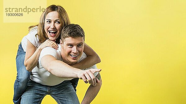 Mann  der seine lachende Freundin Huckepack nimmt und dabei die Kamera auf einen gelben Hintergrund richtet