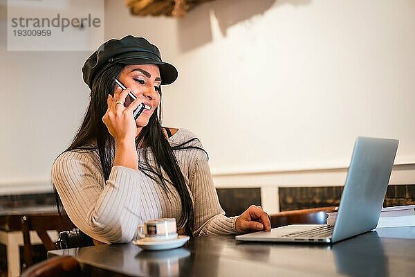 Lächelnde brünette Latina Architektin  die von einem Café aus einen Anruf mit einem Laptop tätigt