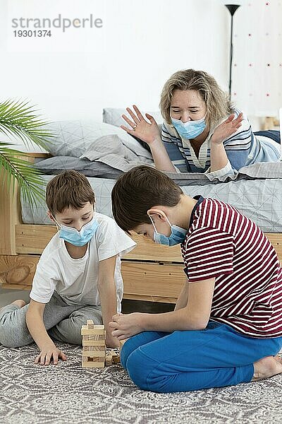 Kinder mit medizinischen Masken spielen zu Hause mit ihrer Mutter Jenga