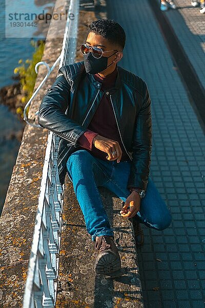 Mode Lebensstil  Porträt eines jungen Latino in den Fluss der Stadt. Jeans  Lederjacke und braune Schuhe. In einer Pandemie mit einer Maske