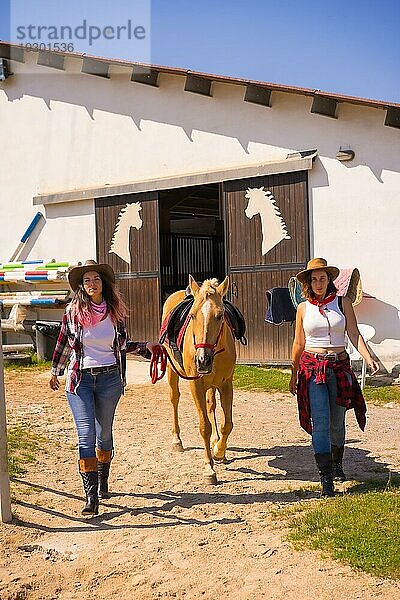 Zwei Cowgirl Frauen kommen lächelnd heraus und ziehen ein Pferd aus einem Stall  mit südamerikanischer Kleidung  Lebensstil von zwei  die in einem Stall arbeiten