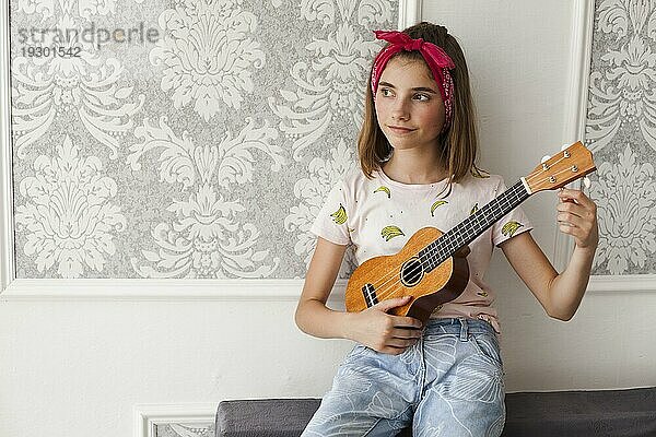 Lächelndes Mädchen  das auf dem Sofa sitzt und die Ukulele einstellt  denkt