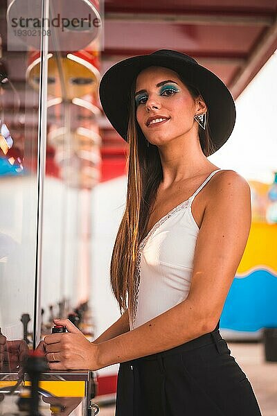 Lächeln einer jungen Brünetten mit schwarzem Hut und weißem TShirt die sich in einem Vergnügungspark an einem Spielautomaten im Sommer vergnügt