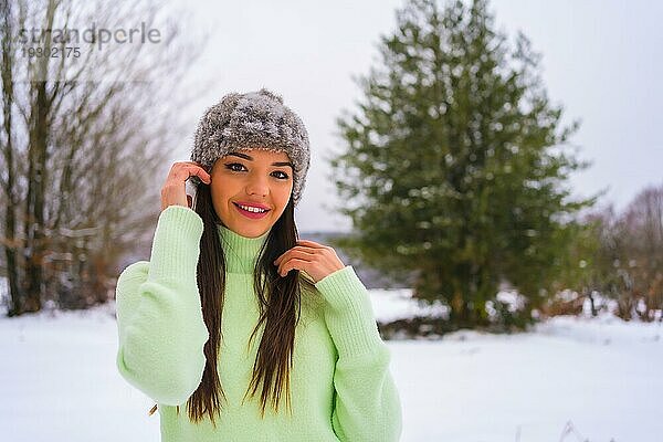 Winter Lifestyle  Porträt junge Brünette Kaukasierin mit grünem Outfit und Wollmütze  lächelnd im Schnee neben verschneiten Kiefern  Urlaub in der Natur