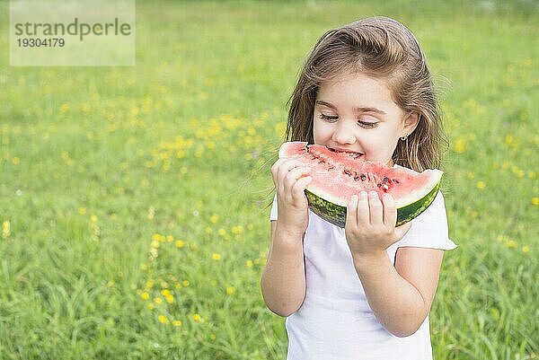 Kleines Mädchen steht im Feld und ißt eine rote Scheibe Wassermelone