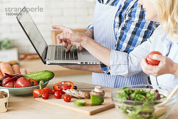 Nahaufnahme einer älteren Frau  die auf einen von ihrem Mann gehaltenen Laptop zeigt  während sie einen Gemüsesalat zubereitet