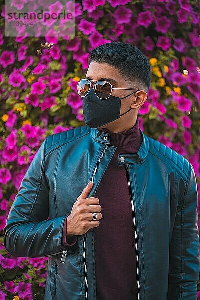 ModeLifestyle Porträt eines jungen Latino mit rosa Blumen im Hintergrund. Jeans  Lederjacke und braune Schuhe. Porträt in der Pandemie mit Maske