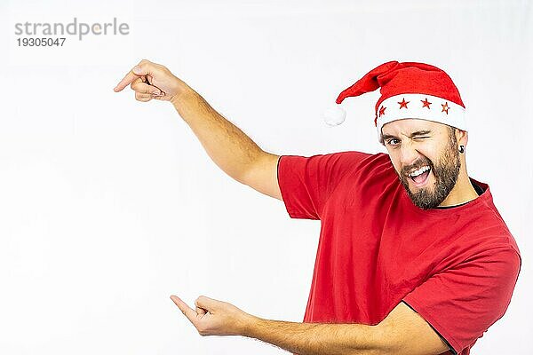 Junger  sehr glücklicher kaukasischer Mann mit roter Weihnachtsmütze  nach links zeigend  auf weißem Hintergrund  bekleidet mit rotem TShirt copy paste