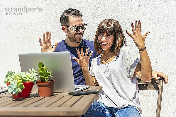 Ein Paar  das zu Hause einen Videogespräch mit einigen Freunden mit dem Computer  auf der Terrasse ihres Hauses mit einem weißen Hintergrund sitzen an einem Tisch. Gruß sehr glücklich