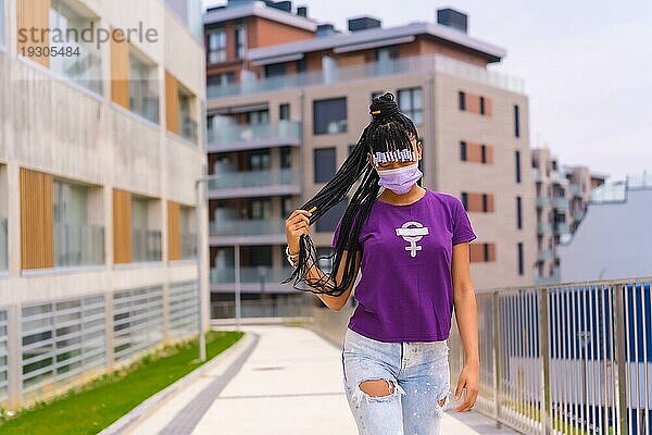 Internationaler Frauentag im Jahr der Coronaviruspandemie Covid 19. Dominikanische Frau mit Zöpfen in einem feministischen lila Hemd und Gesichtsmaske geht durch die Stadt