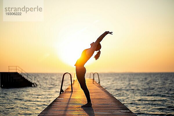 Silhouette einer Frau  die bei Sonnenuntergang auf einem Holzsteg Yoga praktiziert