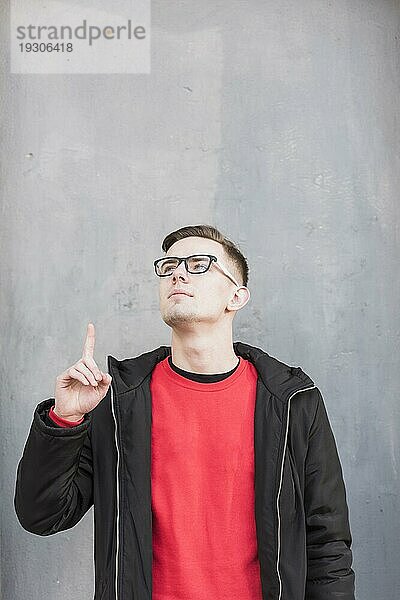 Hübscher junger Mann zeigt mit dem Finger nach oben und schaut gegen eine Betonwand