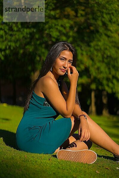 Eine hübsche junge brünette Latina mit langen glatten Haaren  die ein enges grünes Kleid trägt. Porträt eines sitzenden auf dem Gras in einem Park im Sommer