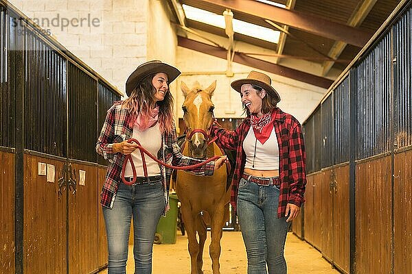 Zwei Cowgirl Frauen haben Spaß und lachen mit einem Pferd in einem Stall  mit südamerikanischen Outfits