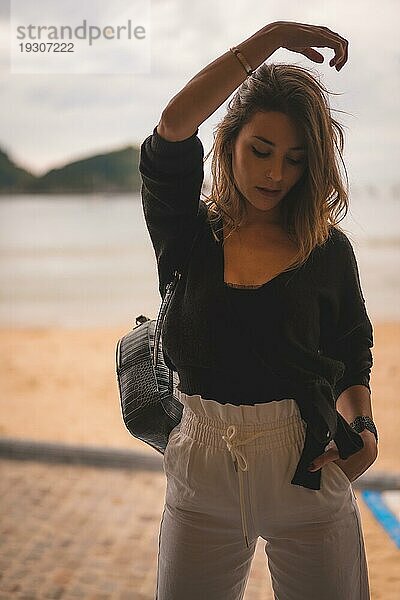 Lifestyle in der Stadt mit einem blonden Mädchen in weißen Hosen und einer Lederjacke in der Nähe des Strandes. Sehr glücklich in einigen Untiefen am Strand in einem Sonnenuntergang