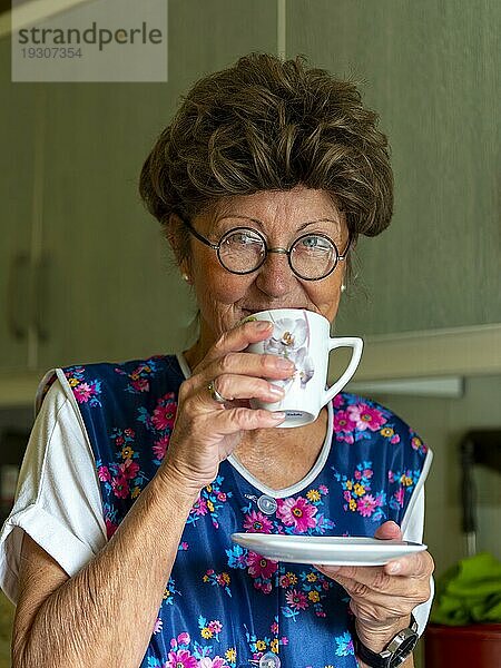 Oma mit alter Kittelschürze  Brille und Perücke in der Küche  hält Kaffeetasse in Händen  Deutschland  Europa