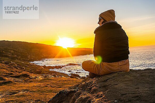 Eine junge Frau mit einer Wollmütze beobachtet im Winter entspannt den Sonnenuntergang auf dem Berg Jaizkibel in der Stadt Pasajes  Gipuzkoa. Baskenland