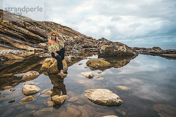 Kaukasisches blondes Mädchen in einem geblümten Hemd  schwarzen Shorts und Strohhut in einer natürlichen Landschaft am Meer und Felsen bei Sonnenuntergang  Lifestyle. Sitzen auf einem Felsen in einem natürlichen Pool