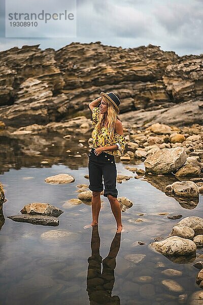 Kaukasisches blondes Mädchen in einem geblümten Hemd  schwarzen Shorts und Strohhut in einer natürlichen Landschaft am Meer bei Sonnenuntergang  Lifestyle. Versteckt in das Wasser eines natürlichen See