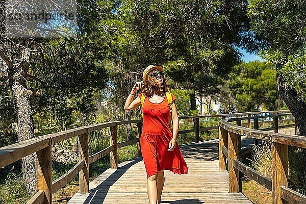 Eine junge Frau in einem roten Kleid geht auf dem Holzsteg in Richtung Strand Moncayo in Guardamar del Segura  Alicante. Gemeinschaft Valencia. Spanien