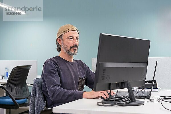 Porträt eines ernsten  stilvollen  reifen Mannes  der an einem Schreibtisch sitzt und einen Computer in einer Arbeitsgruppe benutzt
