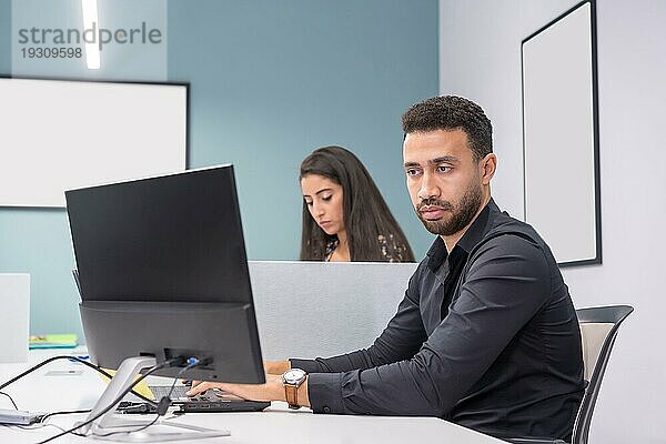 Porträt eines brünetten  modernen und konzentrierten Mannes  der einen Computer in einer Arbeitsgruppe neben einer Frau benutzt