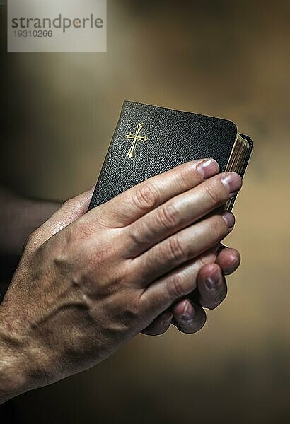 Mann hält eine alte kleine schwarze Bibel in den Händen. Kurze Schärfentiefe  die Schärfe liegt im Kreuz