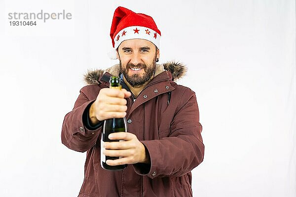 Junger  sehr glücklicher kaukasischer Mann mit roter Weihnachtsmütze  der eine Flasche Champagner auf weißem Hintergrund öffnet  bekleidet mit einer Winterjacke