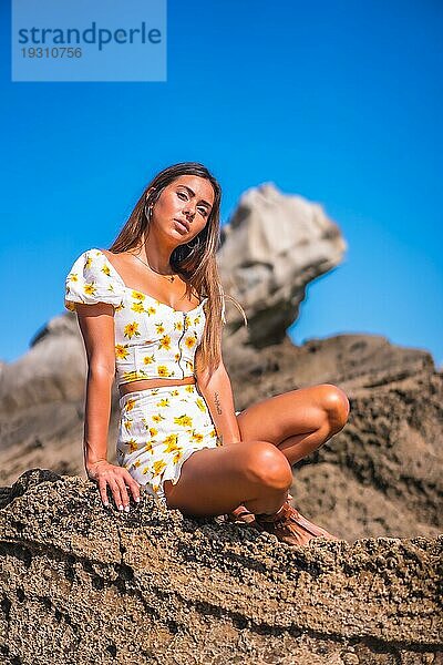 Brünette Modell in einem weißen Kleid am Strand genießen den Sommer  sitzen auf einigen Felsen